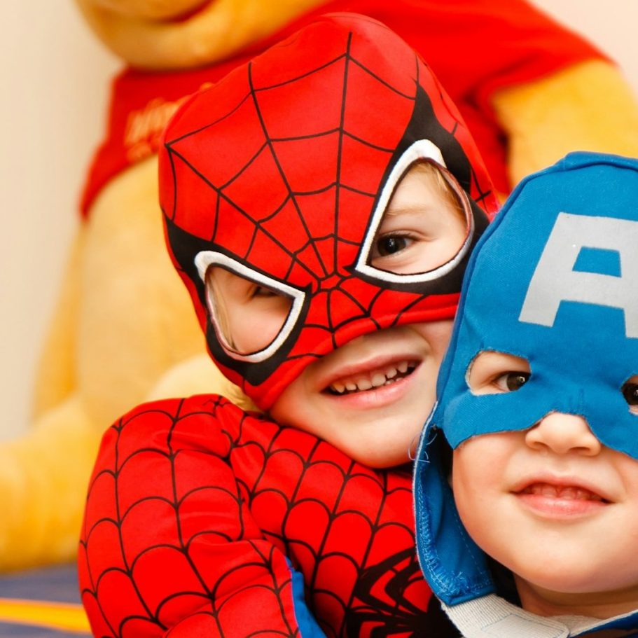 Kinder im Spiderman Kostüm Kinder in Superhelden-Kostümen, lächelnd, hinter einem gelben Teddybär.