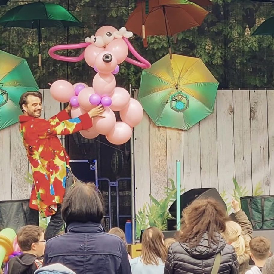 Kinderzauberer Maik bei seiner Bühnenshow Kinderzauberer in buntem Kostüm erschafft einen rosa Ballontierhund vor einer Gruppe Kinder.